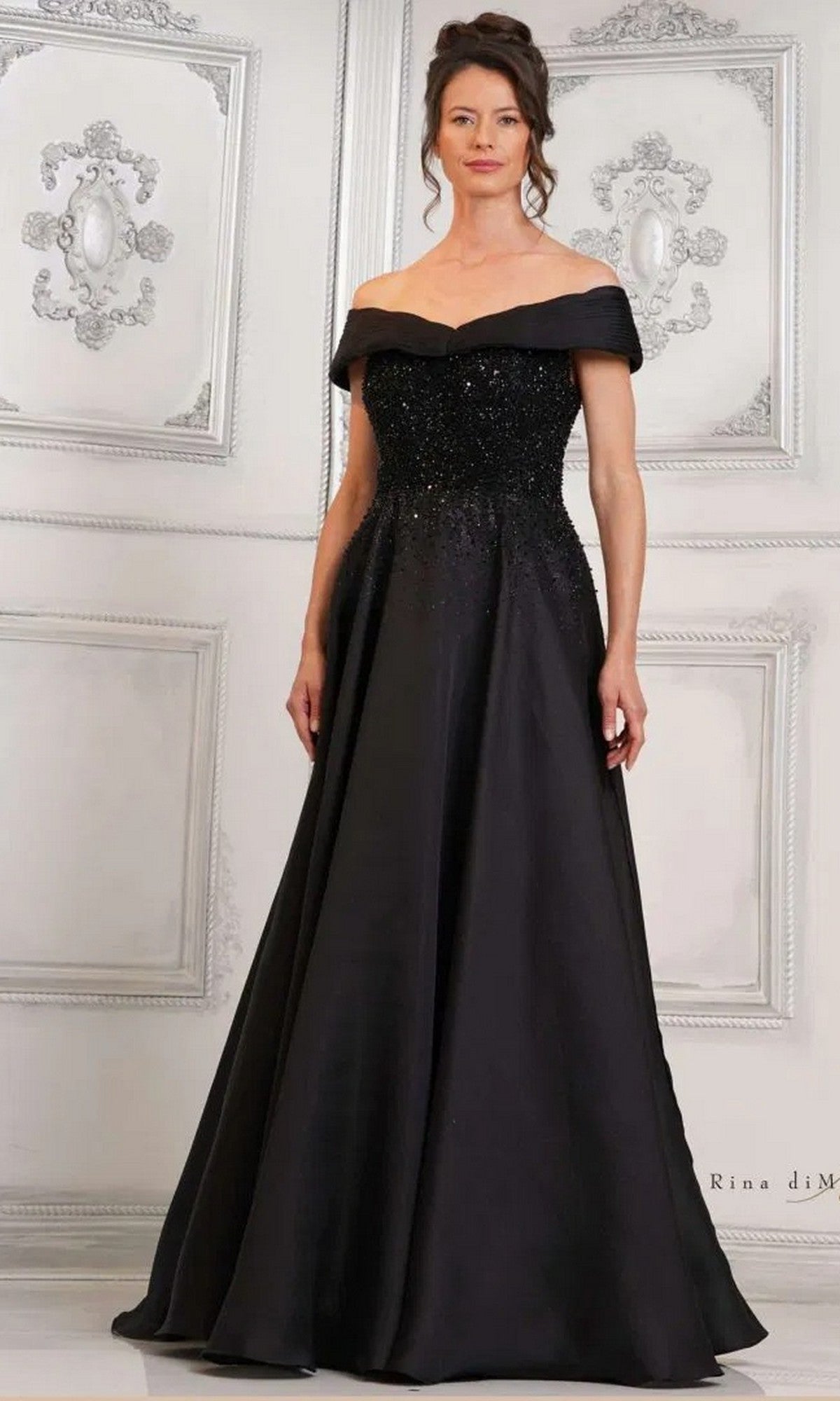 A woman poses against an ornate white wall in the black Rina Di Montella RD3102 long formal dress, featuring an off-the-shoulder beaded bodice and a floor-length A-line skirt.
